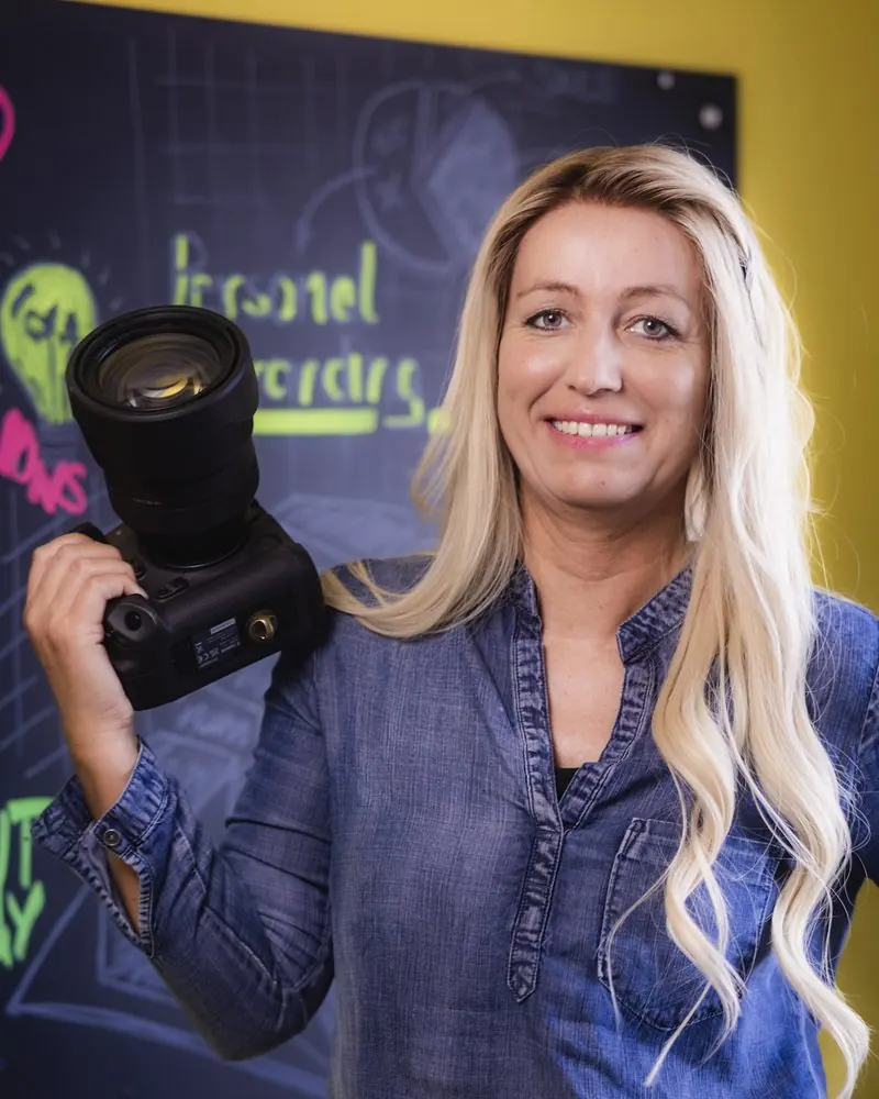 Portrait einer Fotografin mit Kamera vor einer bunt beschrifteten Tafel