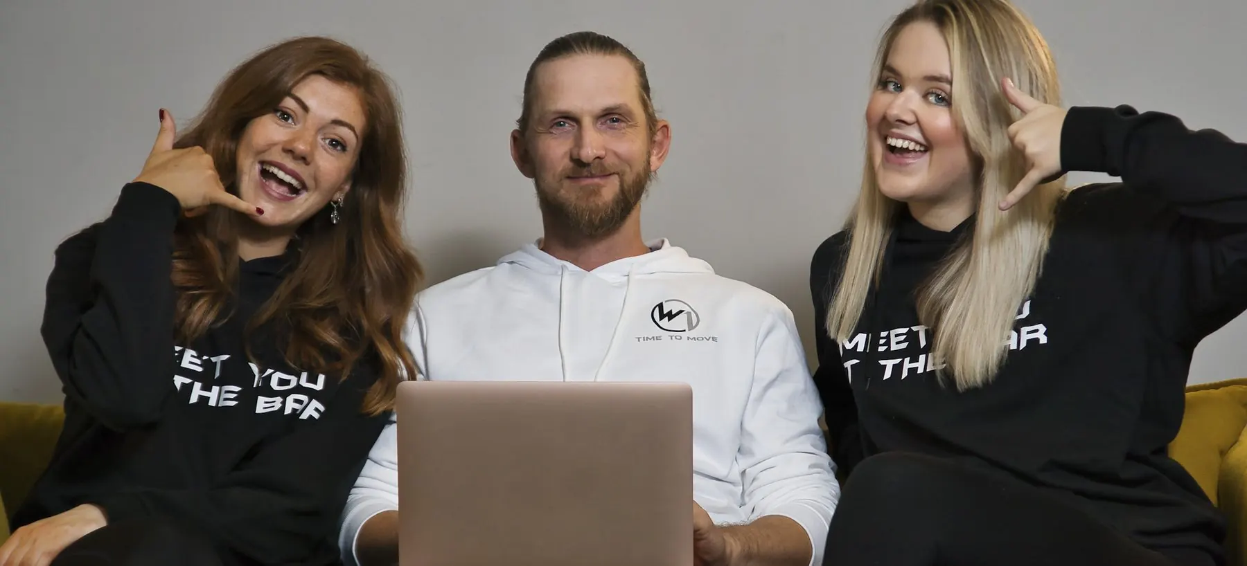 Teamfoto: drei Mitarbeitende sitzen gut gelaunt mit einem Laptop auf einem gelben Sofa